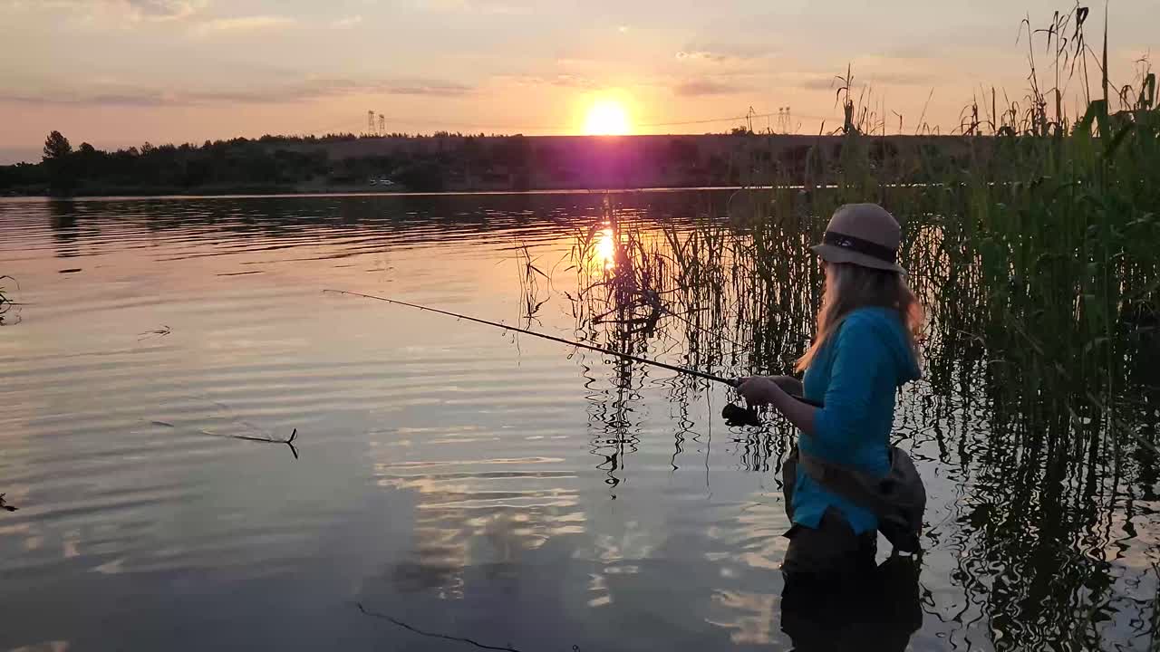A woman fishing and wading in a tranquil lake with a stunning sunset in the background