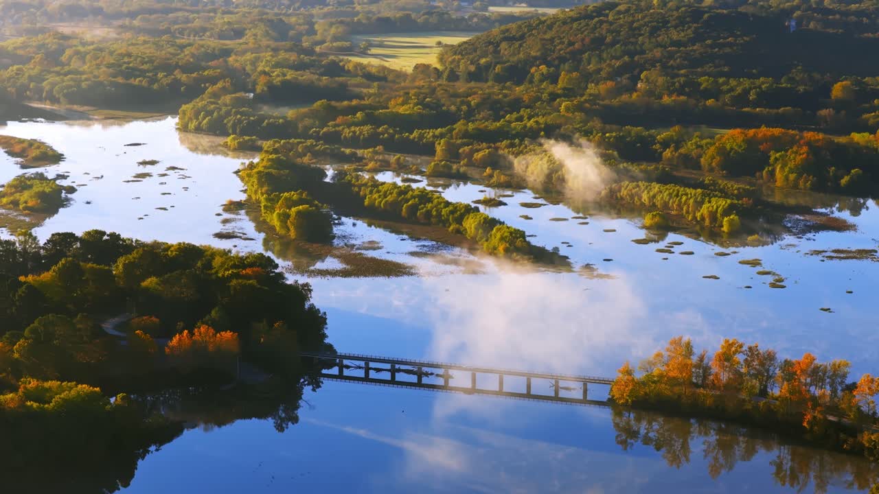 Air biru jernih yang dibatasi oleh garis pantai musim gugur yang semarak, dengan jembatan yang terhubung di Danau Sequoyah, terbang secara aerial menembus awan berkabut, Wyman Arkansas AS.