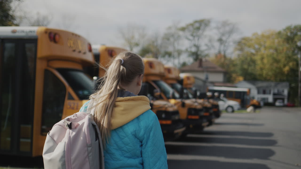 En elev går langs en række gule skolebusser. Bagfra-visning.