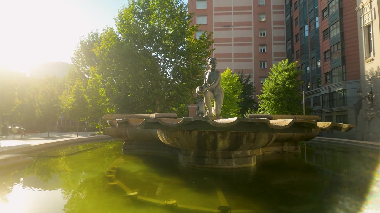 Una serena fuente de piedra con una estatua femenina se alza en una tranquila plaza de Madrid durante una tarde soleada, capturando el auténtico bienestar urbano y la sostenibilidad en España.