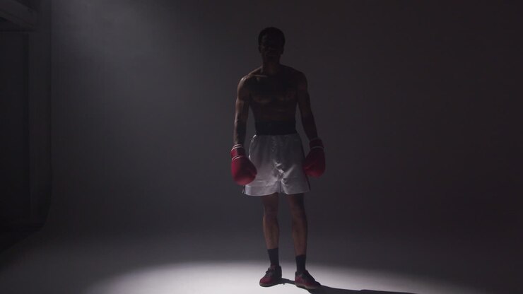 Un boxeador posa en un estudio con un foco sobre él, mostrando sus músculos y guantes de boxeo