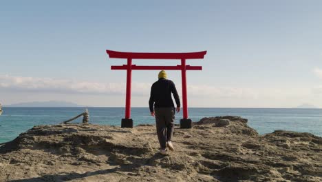 Varón-Joven-Caminando-Hacia-La-Puerta-Torii-Roja-Japonesa-Frente-Al-Océano