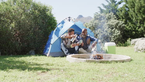 Happy-african-american-father-and-adult-son-sitting-outside-tent-drinking-coffee-in-sun,-slow-motion