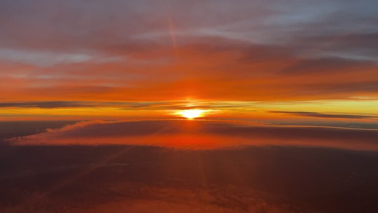 Una coinvolgente visuale in prima persona dalla cabina di pilotaggio di un jet che vola tranquillamente al tramonto tra strati di eteree nuvole color arancio, con il sole che tramonta dietro l'orizzonte