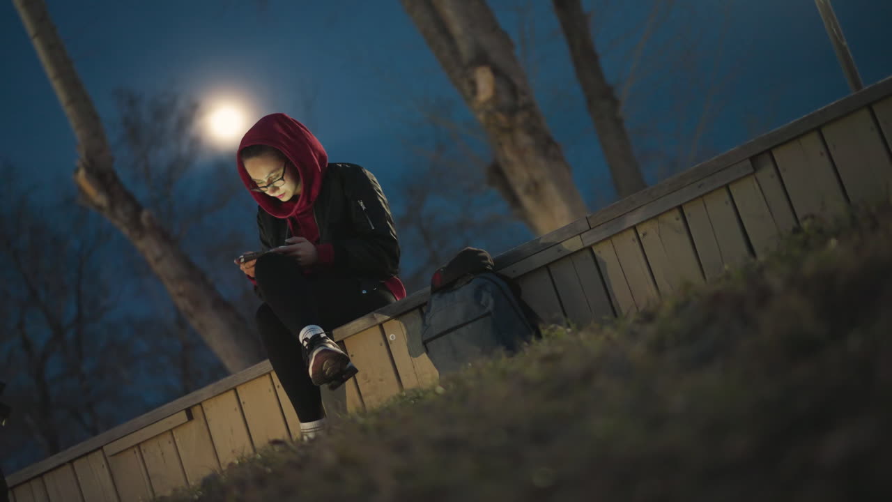 Moonlit Park Bench Lone Teen Texting, Red Hoodie Lit By Phone Glow, Backpack At Side, Bare Trees And Full Moon Halo, Quiet Urban Night Mood With Introspective Atmosphere And Subtle Streetlight Shadows