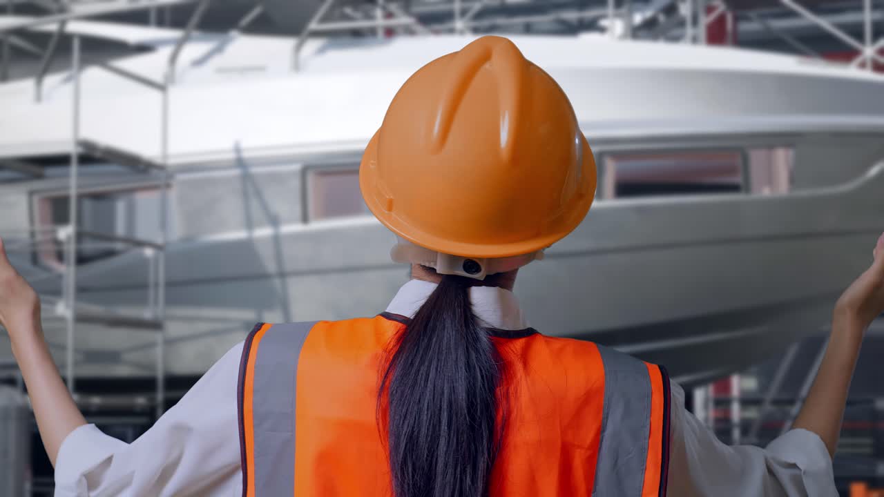 Primo piano, vista posteriore di un'ingegnera con casco di sicurezza che allarga le braccia mentre è in piedi vicino alla costruzione di uno yacht in un moderno cantiere navale