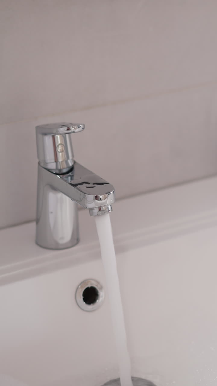 una corriente de agua fluye de la grifa al fregadero en un baño brillante en cámara lenta. la hembra enciende el grifo de agua para lavarse la cara después de la esteticista
