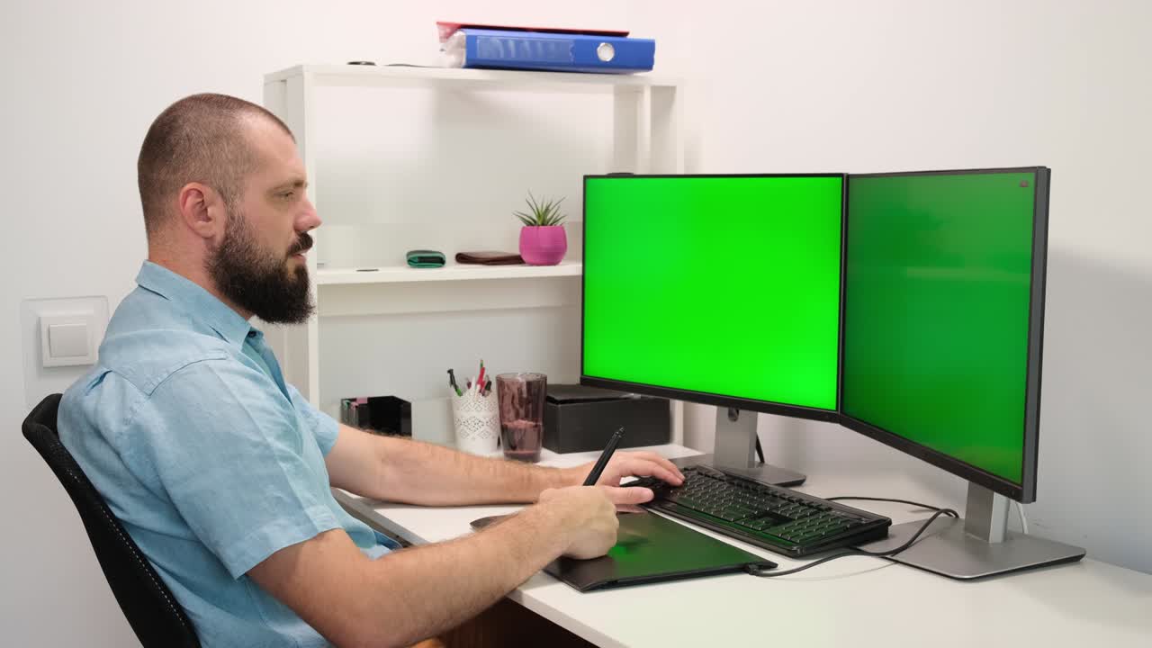 un hombre dibuja en una tableta gráfica. un hombre trabajando en casa frente a dos monitores verdes.