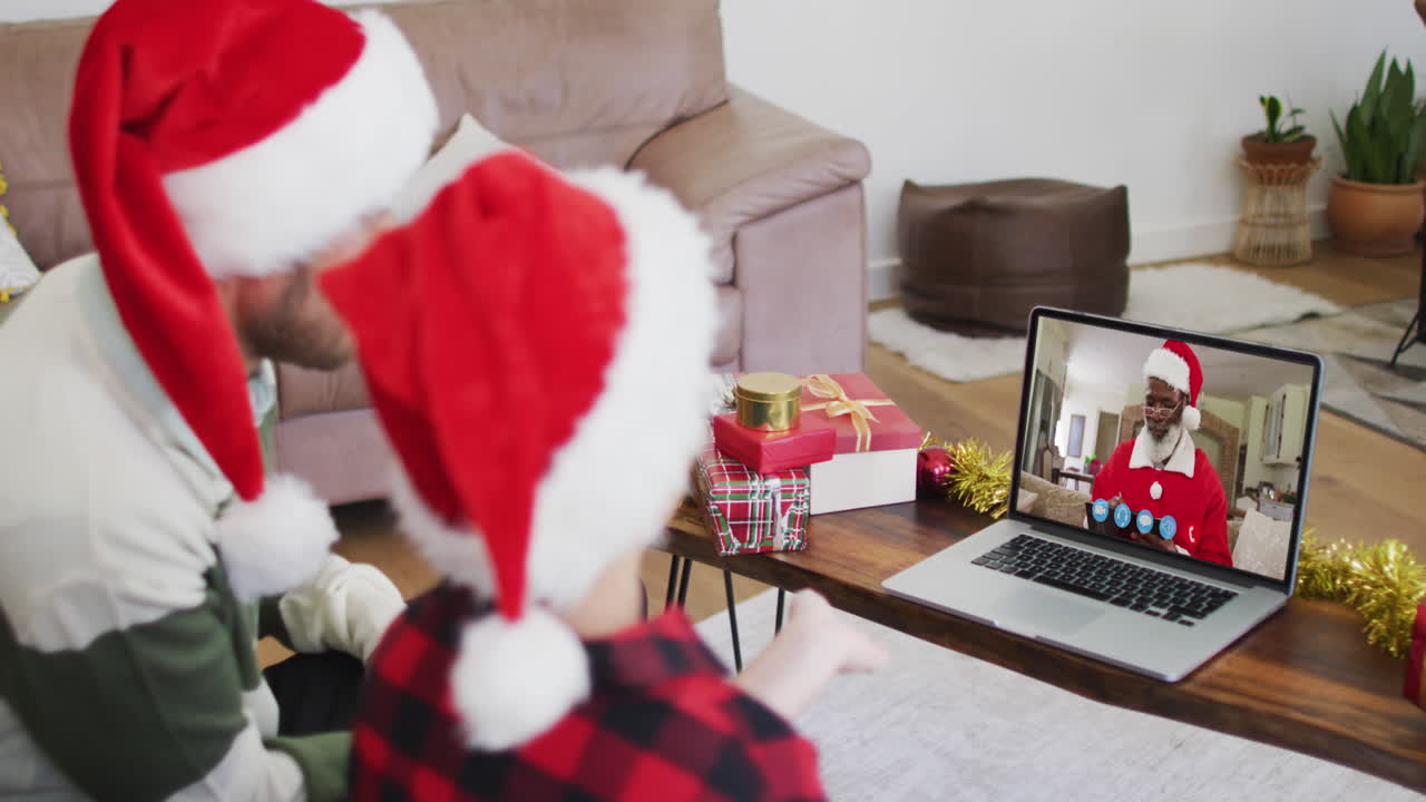 hombre caucásico con hijo usando sombreros de santa en una videochat portátil durante la navidad en casa