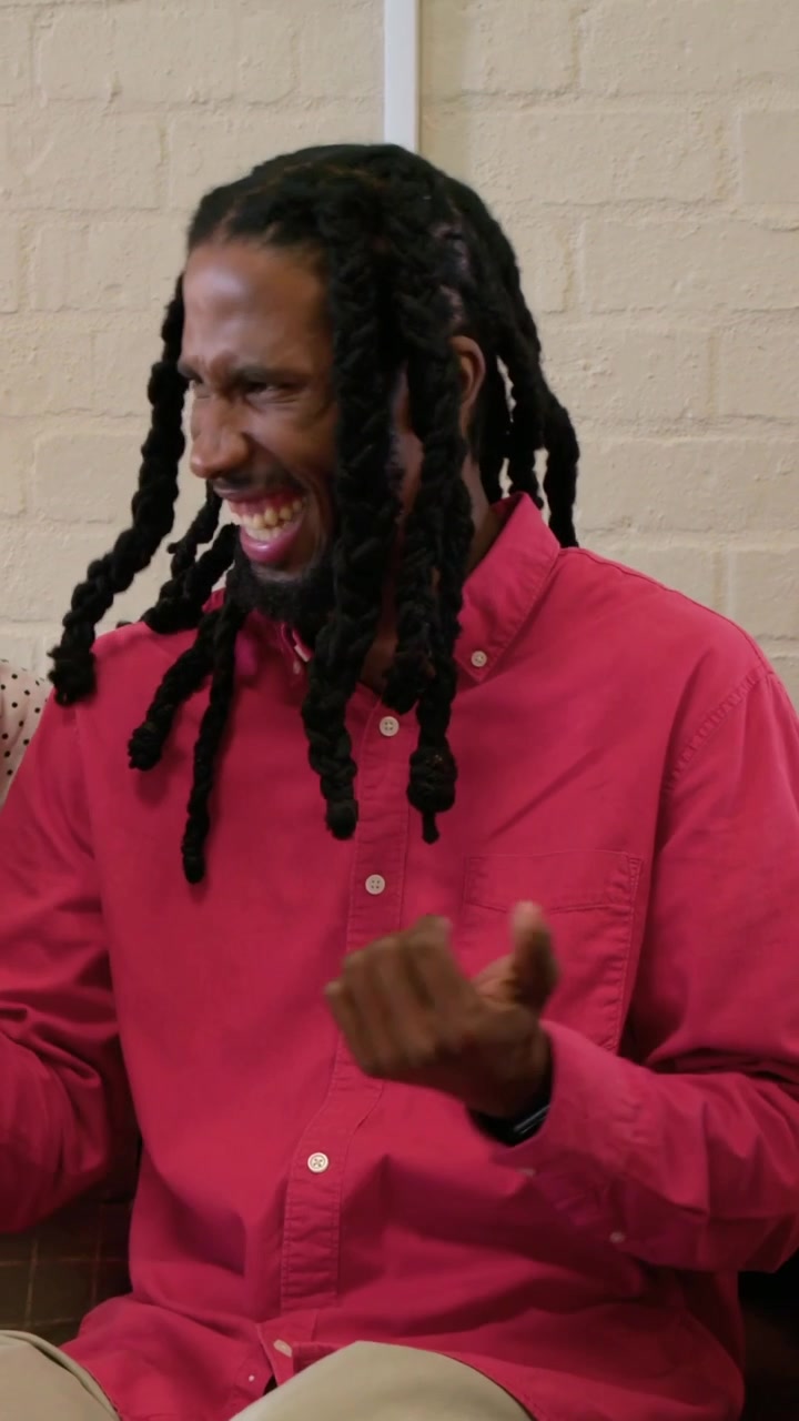 Vertical video: Laughing man in red shirt responding to partner touch on couch, polka-dot blouse