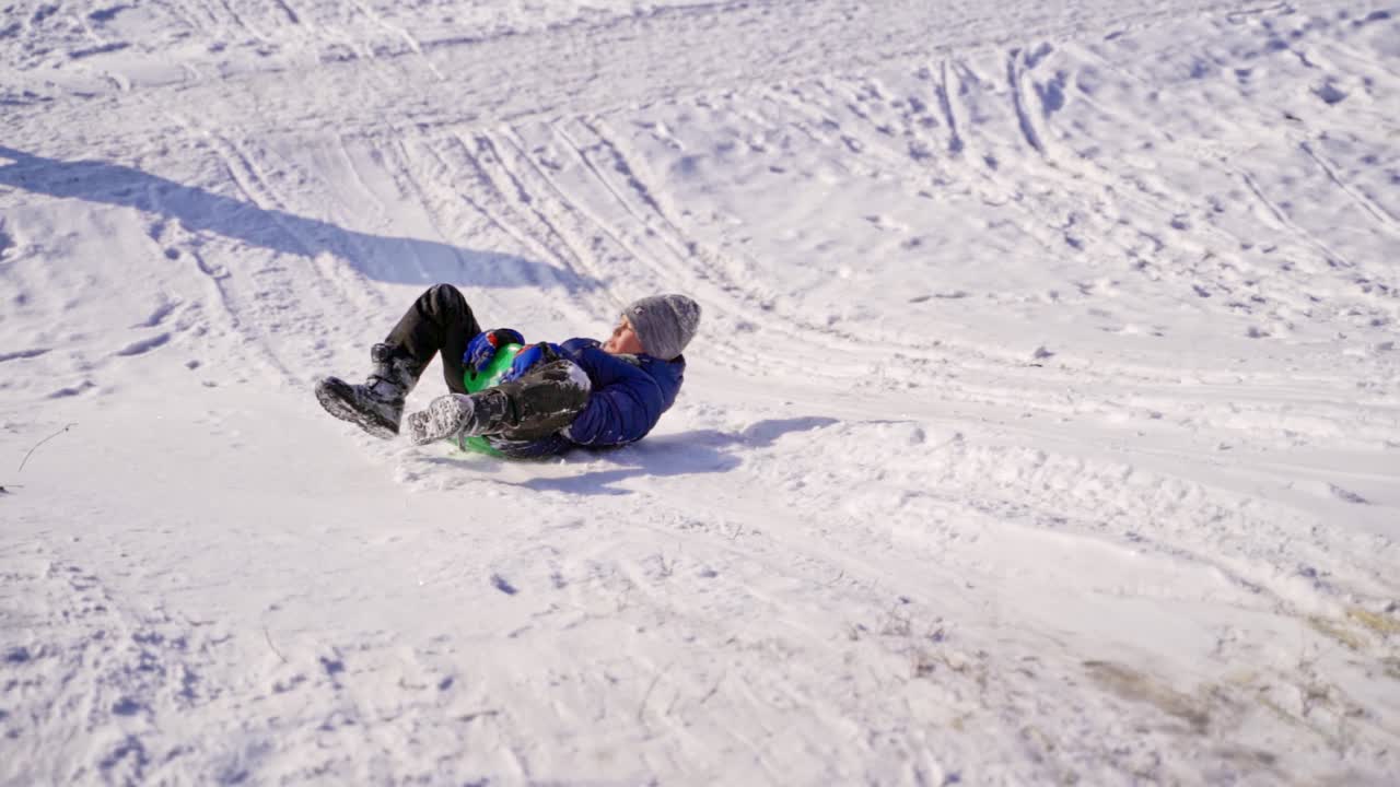 Boy sledding down the hill in winter. Cute boy with saucer sled riding from snowy slide outdoors. Slow motion.