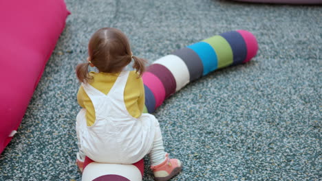 funny 3-year-old little girl pretends to ride on a colorful long pillow in a play area of a shopping mall