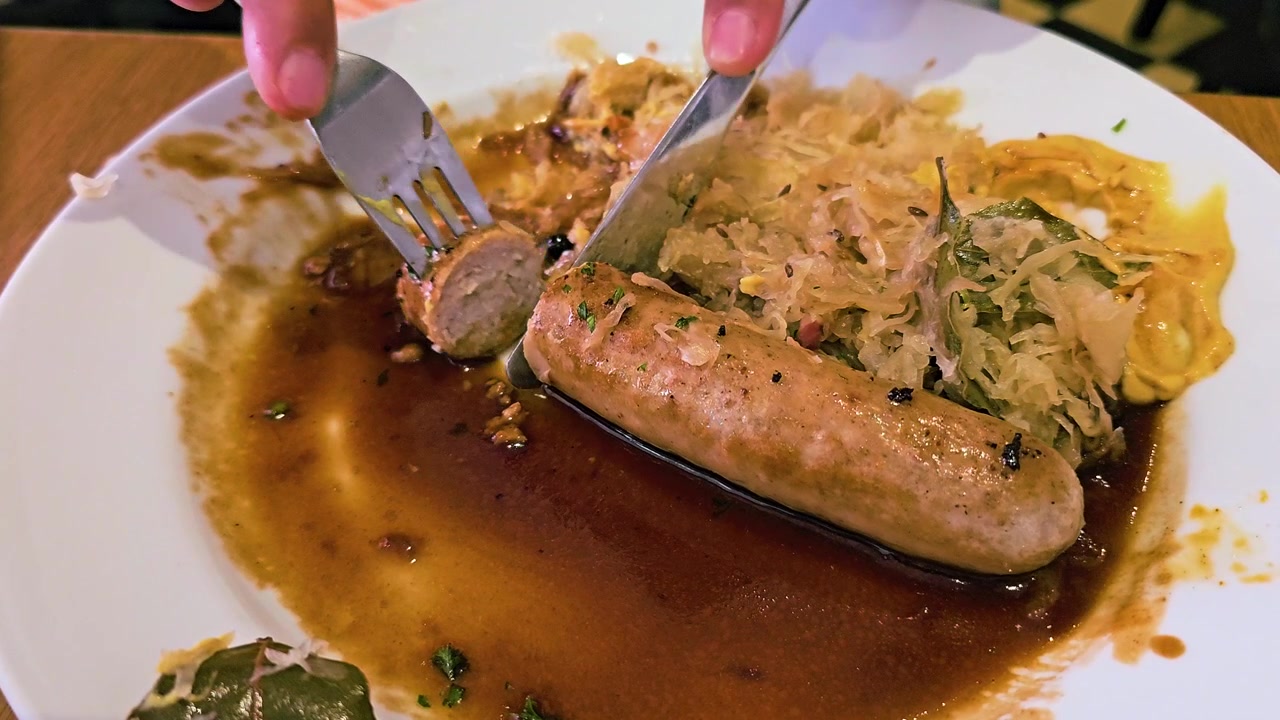 Manos usando tenedor y cuchillo para cortar salchicha bratwurst servida con chucrut, mostaza y una rica salsa marrón, primer plano de abundante cocina alemana en un plato blanco en un acogedor ambiente de restaurante