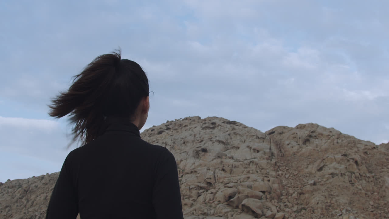 바람이 머리카락을 움직이는 동안 모래 산을 바라보며 사막에서 검은 옷을 입은 신비한 아름다운 여성의 슬로우 모션 샷