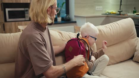 Ein-Glücklicher-Blonder-Mann-Mit-Brille-Und-Bart-Hilft-Seinem-Kleinen-Sohn,-Einem-Albino-Jungen-Mit-Blauer-Brille,-Einen-Rot-orangen-Rucksack-Anzuziehen-Und-Begleitet-Seinen-Kleinen-Sohn-Morgens-Zur-Ersten-Unterrichtsstunde-In-Einer-Modernen-Wohnung