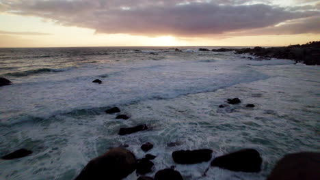 aerial pull-in of waves crashing along the rocky shoreline in cape town, south africa, moving towards the expansive sea and a vibrant, colorful sunset with clouds on the horizon.