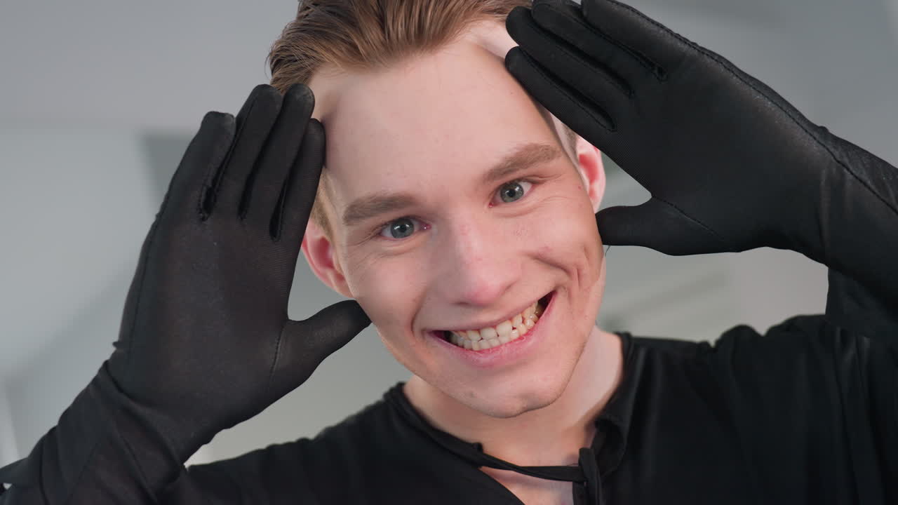 close up of playful young man in black gloves framing face with hands, flashing bright smile and lively eyes in modern mirror studio, conveying energetic fun and confident expression