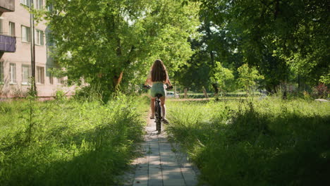 back view of young woman confidently riding bicycle through a lush green park path, her long hair flows freely, blending with natural scenery, background includes trees and a building