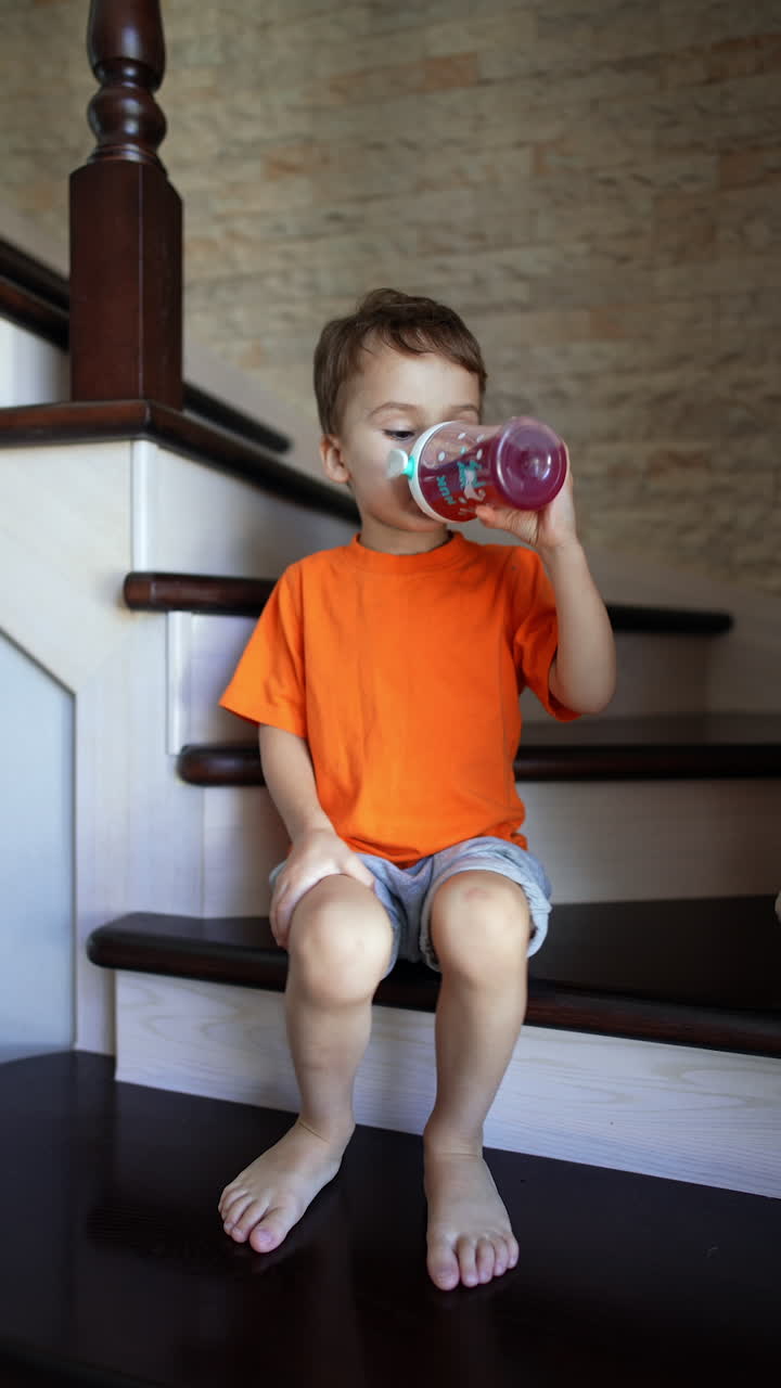 Caucasian toddler sitting on the stairs at home drinks compote. Cute kid puts the bottle and stands up to leave. Vertical video.