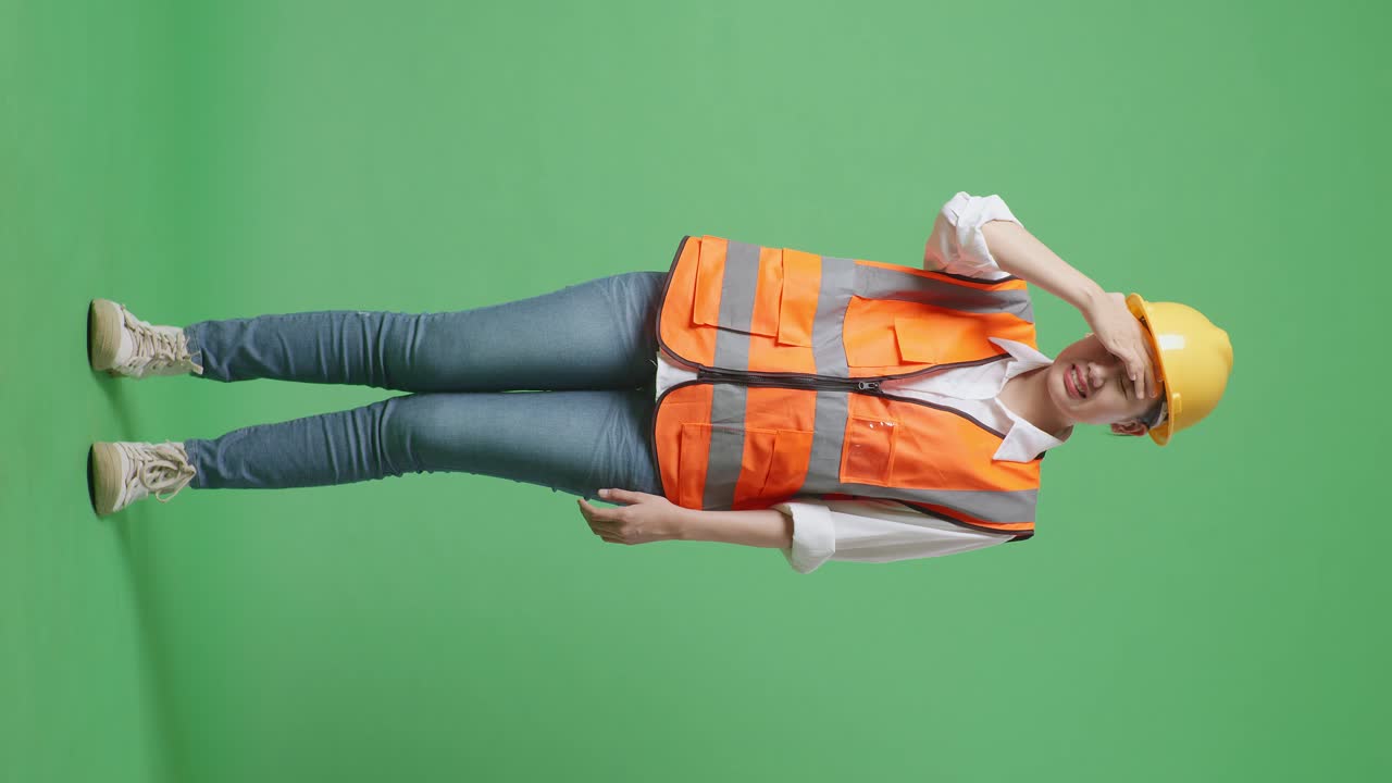 안전 헬을 입은 아시아 여성 엔지니어가 녹색 스크린 배경 스튜디오에서 일하는 동안 두통을 고 있습니다.