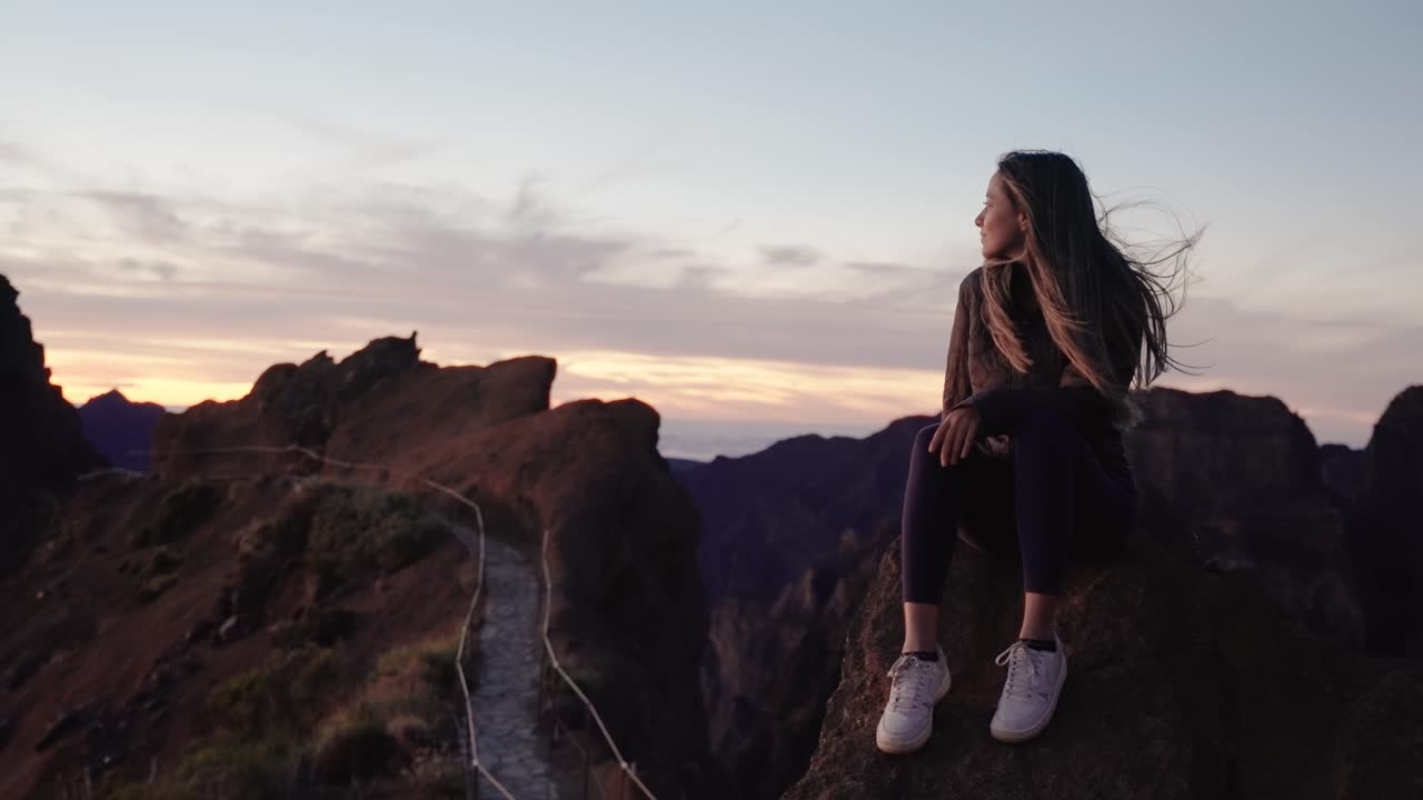 A woman sits on a rocky cliff edge with her hair blowing in the wind, watching the sunrise in Madeira, Portugal.