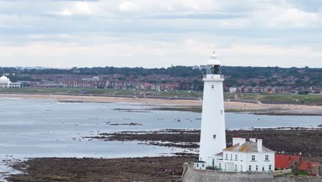 a steady horizontal pan reveals a tall white lighthouse beside rocky tidal flats, with distant houses under overcast daylight. smooth camera movement, wide landscape view