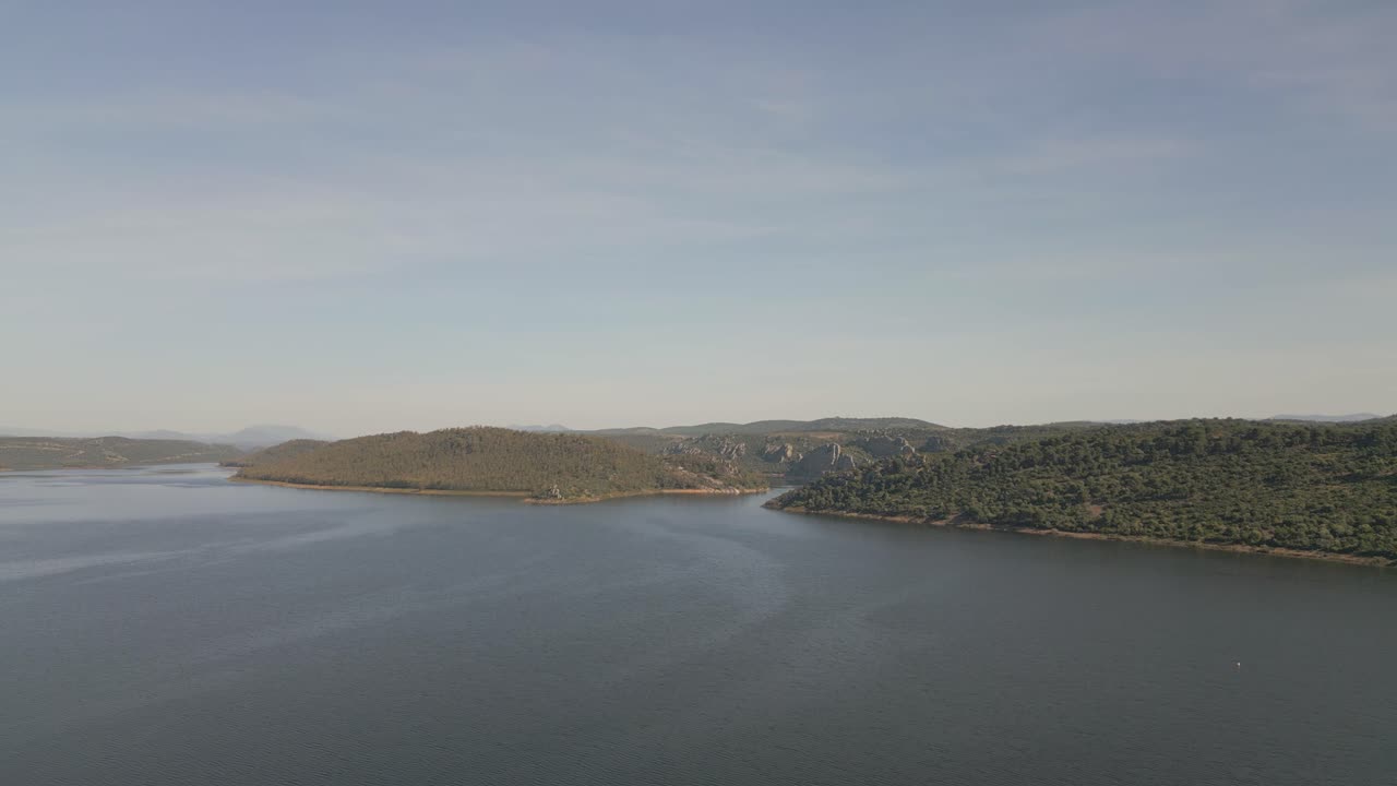 Aerial footage of Cíjara Reservoir in Badajoz, Spain, showing calm blue waters and the rocky hills rising on the horizon under a sunny sky. A stunning natural summer landscape of serenity and beauty