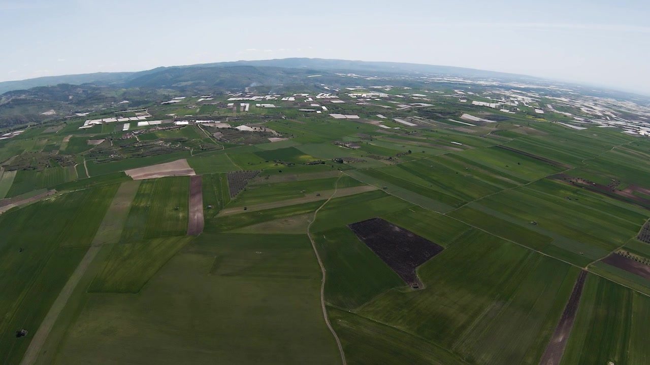 FPV drone shot over the flat patchwork farmland of the Sicilian interior, with greenhouses, cultivated fields and distant mountains stretching to the horizon under a wide open sky.