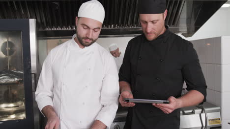 focused cucasian male chef instructing trainee male chef using tablet in kitchen, slow motion