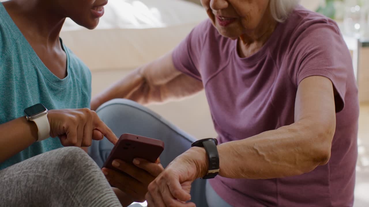 African American woman caregiver pointing at phone, guiding senior woman checking tracker at home