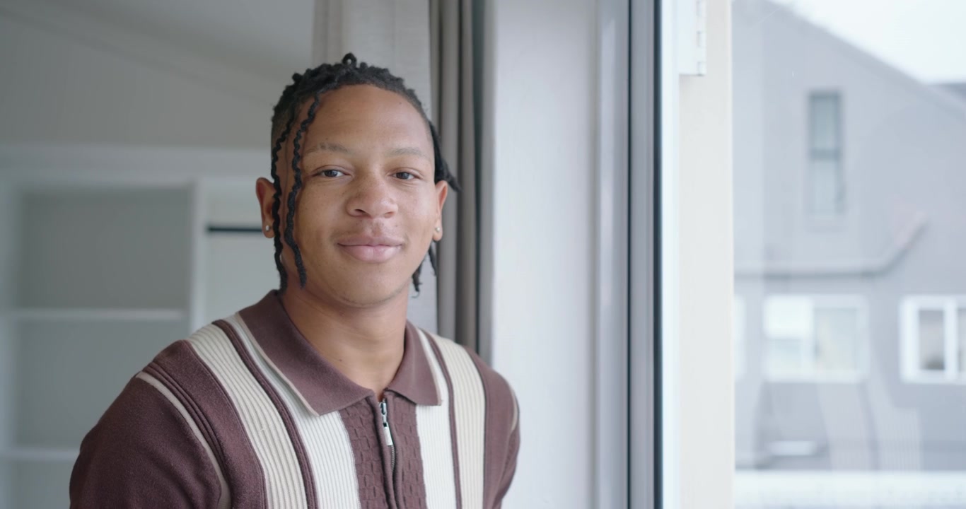 Uomo afroamericano che indossa una felpa con zip a righe alla finestra, girandosi verso la telecamera che si avvicina, sorridendo.