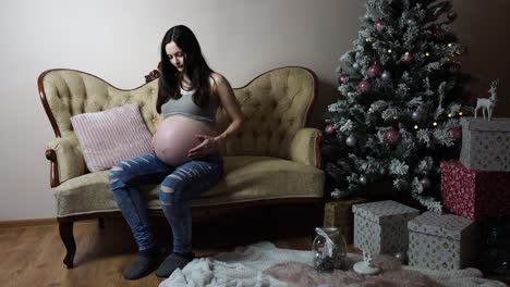 mujer embarazada se sienta cerca del árbol de navidad y toca suavemente su gran vientre redondo
