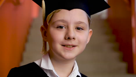 Retrato-De-Un-Feliz-Estudiante-De-Preescolar-Con-Toga-Y-Birrete-Sosteniendo-Un-Libro-Y-Mirando-La-Cámara-En-La-Ceremonia-De-Graduación