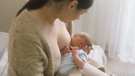 自宅のベッドに座りながら生まれたばかりの赤ちゃんに母乳を与える愛情のある若い母親1