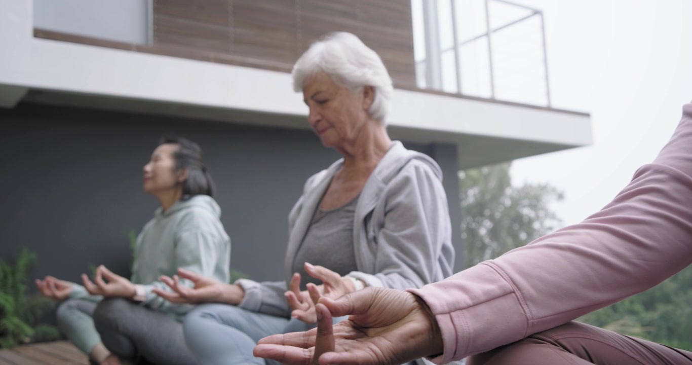 Forskellige kvindelige venner mediterer i skrædderstilling på yogamåtter på et trædæk for at finde ro, mens kameraet panorerer.