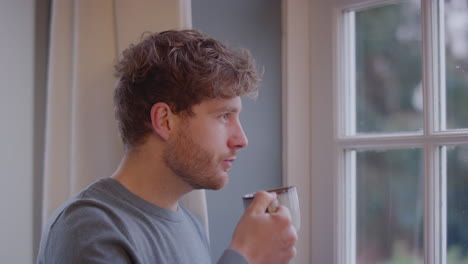 joven en casa pensativo mirando por la ventana y bebiendo una taza de café