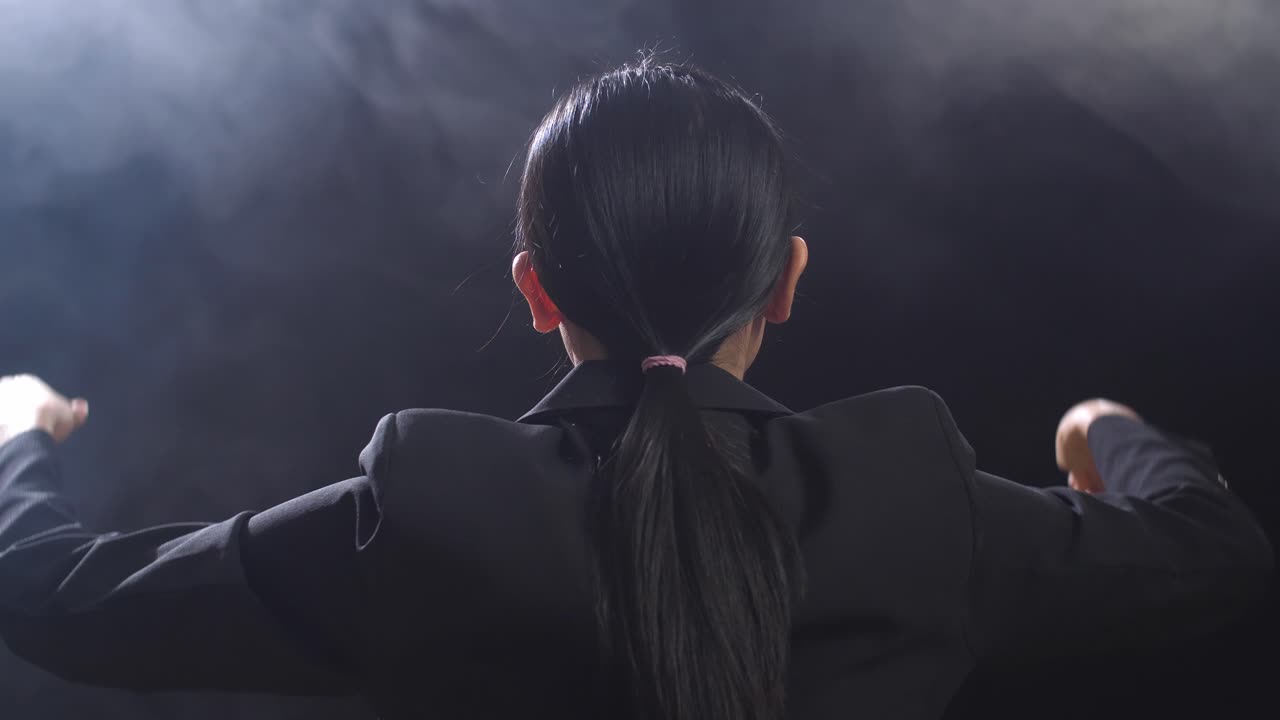 뒷면에서 가까이 쳐다보는 아시아의 지휘자 여성은 막대기를 들고 안개가 있는 검은 스튜디오에서 빠르게 몸짓을 보여줍니다.