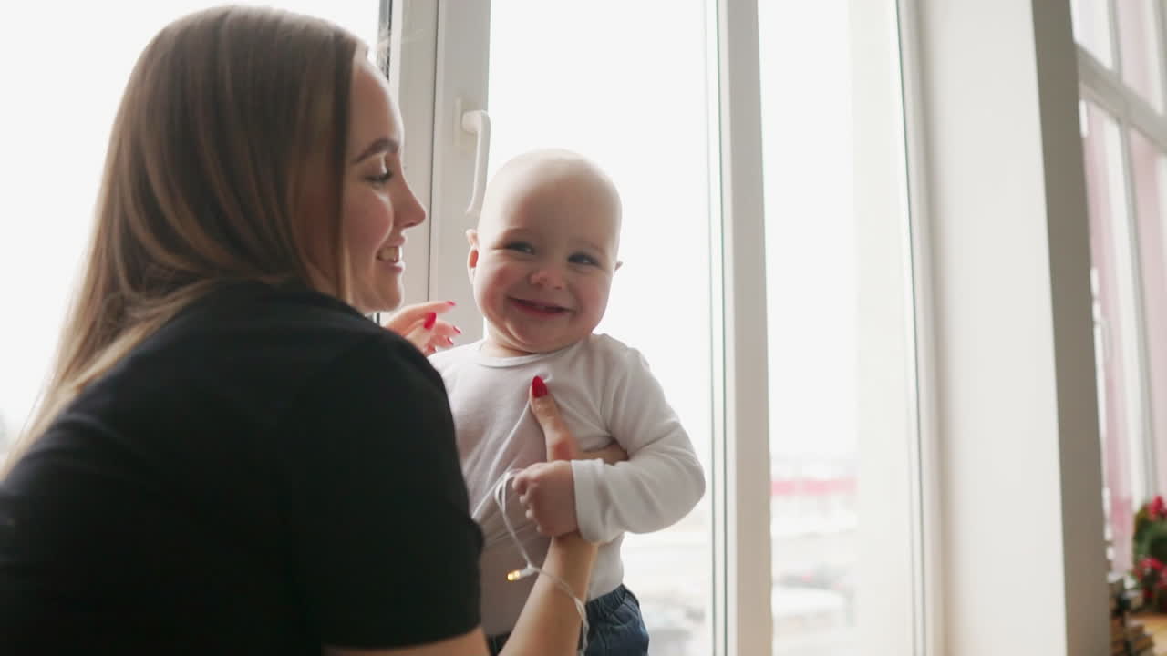 Young mother is holding her son's hands while he is walking on the window sill decorated with Christmas wreath. Happy family kissing