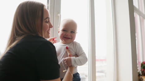 Young-mother-is-holding-her-son's-hands-while-he-is-walking-on-the-window-sill-decorated-with-Christmas-wreath.-Happy-family-kissing