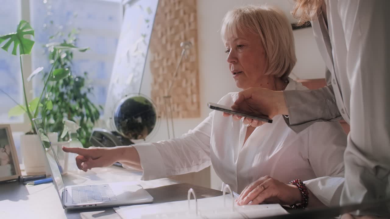 A woman working at a desk with a laptop and being assisted with a smartphone