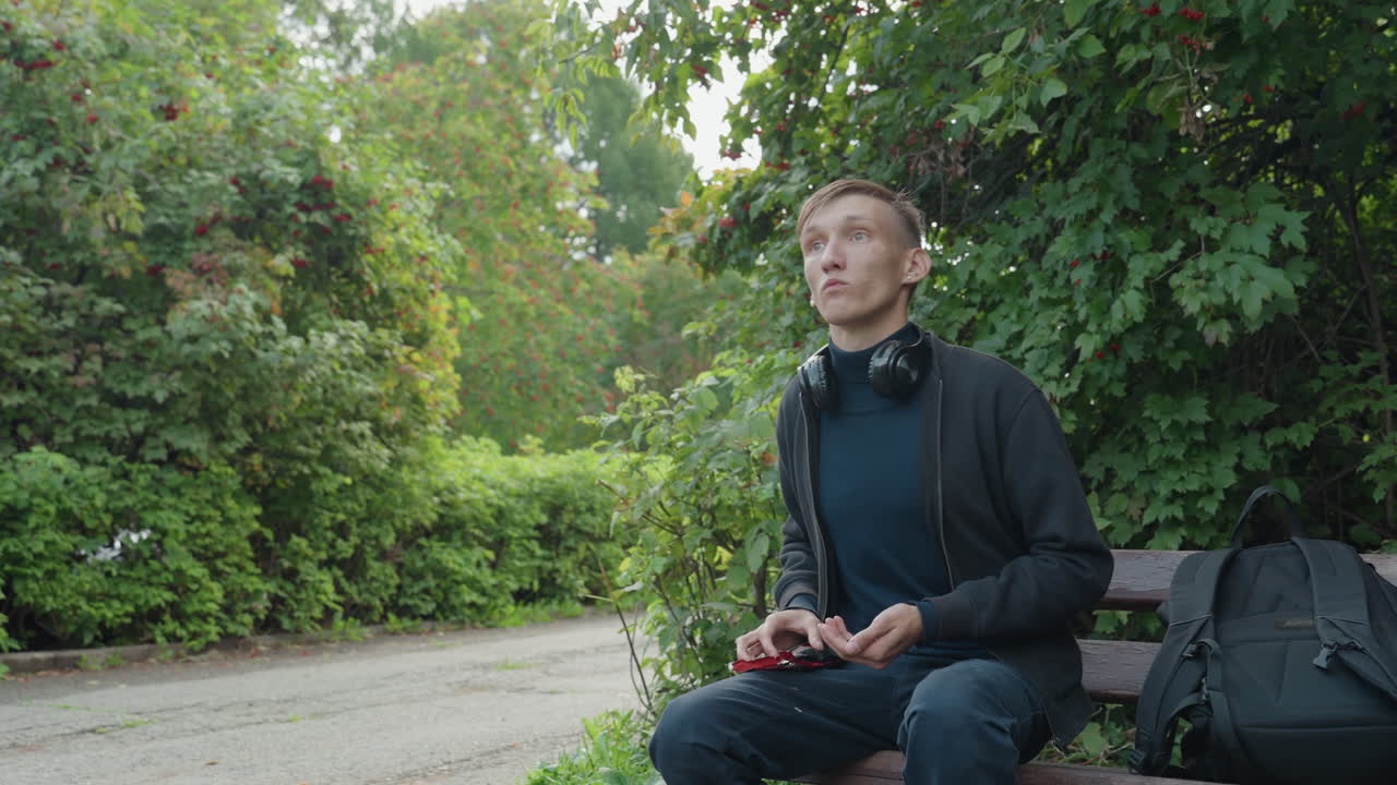 Ung kaukasisk mand sidder på en bænk, scroller på telefonen, justerer hovedtelefonerne ved siden af rygsækken i en grøn park og stopper op for at se opad i et eftertænksomt studieøjeblik med blødt, overskyet lys i en stemning af urban ensomhed.