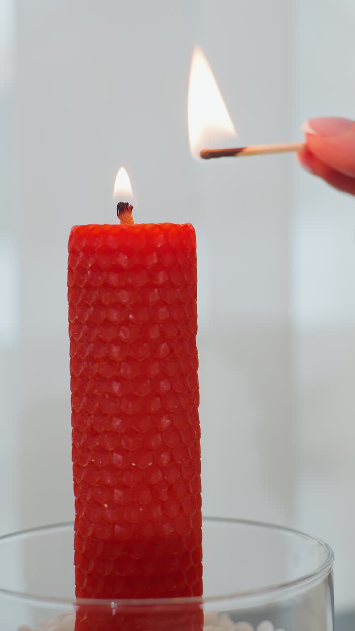 Primo piano di una mano che accende una candela rossa con un fiammifero sul tavolo della cucina; la luce calda crea un'atmosfera accogliente, con burro, piatti e bicchieri sullo sfondo.