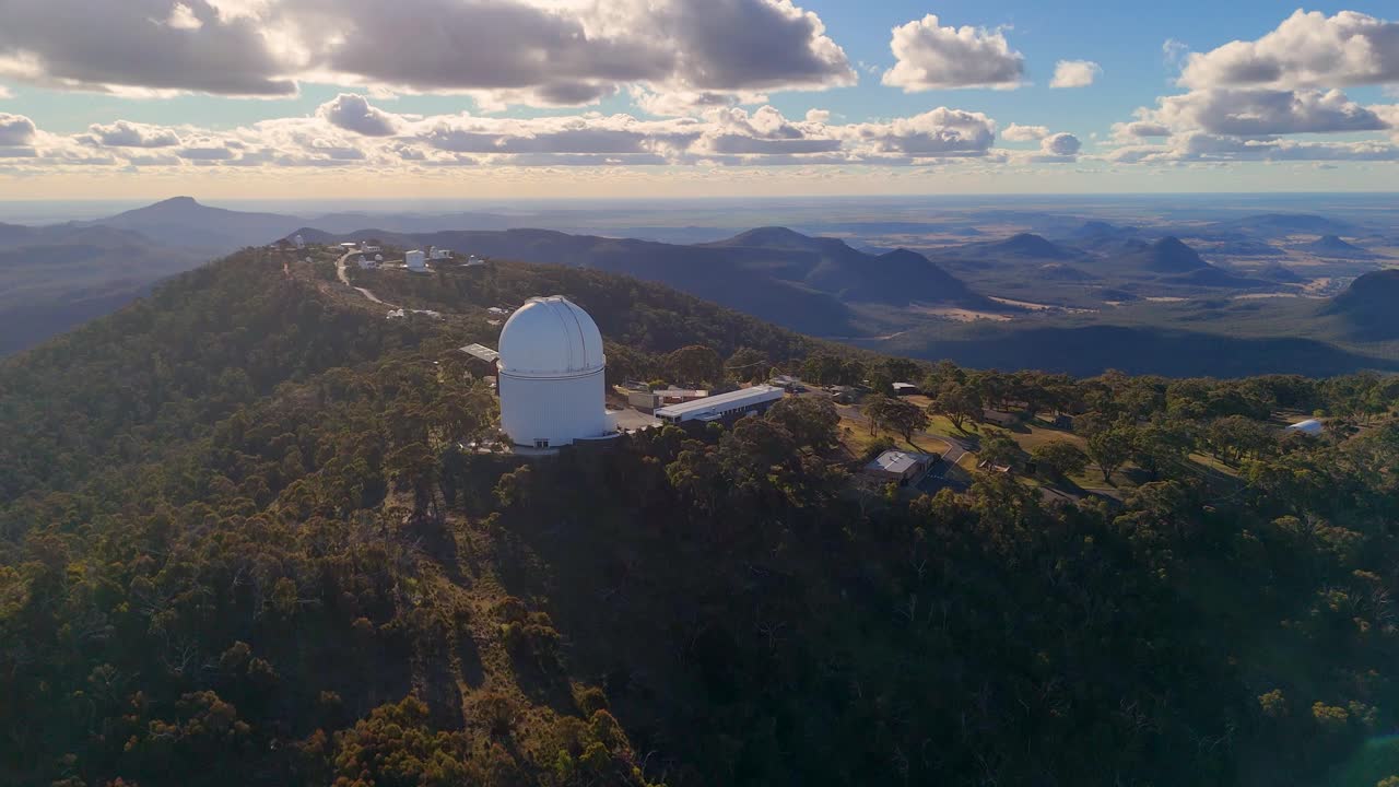 Aerial footage glides over a mountaintop observatory surrounded by forest, with dramatic clouds and warm sunset light illuminating the landscape and distant hills