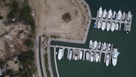 Luftaufnahme-Aus-Der-Vogelperspektive-über-Einen-Yachthafen-Von-Marina-Puerto-Los-Cabos-In-Mexiko-Mit-Blick-Auf-Boote-Und-Yachten,-Die-Auf-Den-Anlegestellen-Im-Ruhigen-Hafenwasser-Angeordnet-Sind,-Und-Auf-Eine-Straße,-Die-Sich-Entlang-Der-Küste-Erstreckt