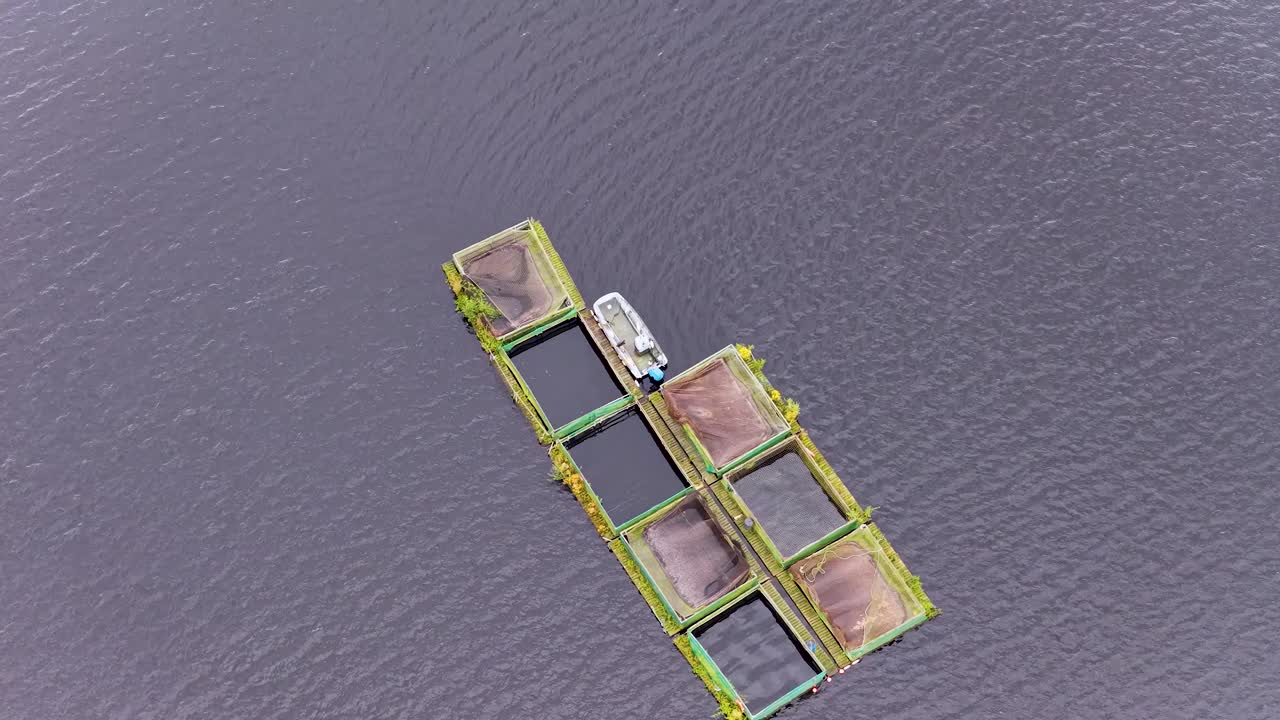 Overhead drone footage shows a boat moored to floating fish cages on calm reservoir water, with steady camera movement and soft natural daylight