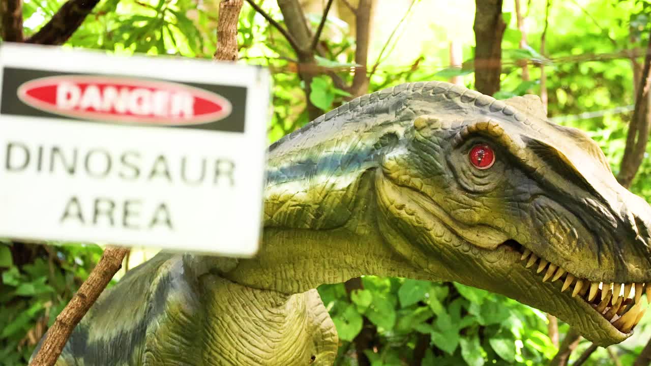 A dinosaur model amidst lush greenery with a warning sign. Bright daylight highlights the scene's vivid colors
