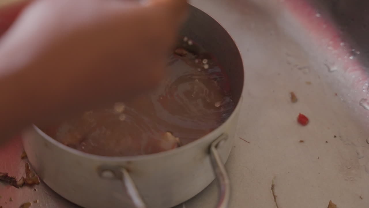 Hands stirring murky water in pot at sink, unspecified cook scrubbing residue with circular motion to clean saucepan soapy bubbles, metal pan, domestic dishwashing chore carried out with steady rhythm