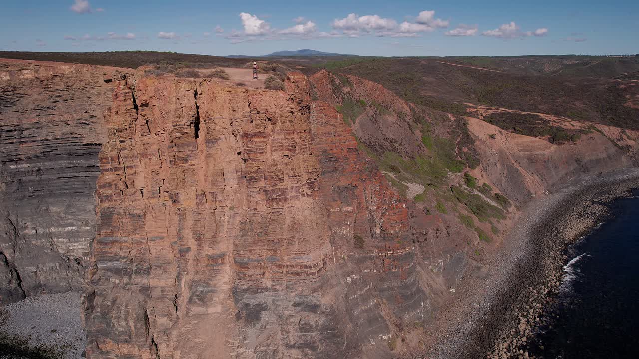 Uomo in piedi su una scogliera di arenaria stratificata a Vila Nova de Mil Fontes, Alentejo, Portogallo, sopra l'Oceano Atlantico, evidenziando l'imponenza della drammatica falesia e il paesaggio di erosione costiera, con drone in orbita.