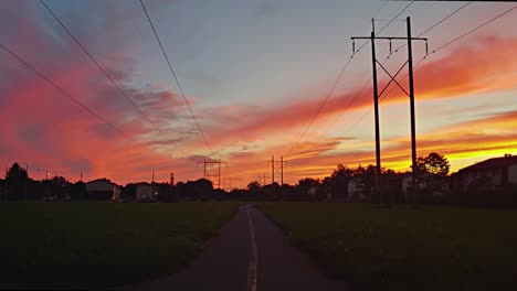 Timelapse-of-a-trail-on-a-summer-evening-with-a-huge-sunset-and-lights-turning-on-as-it-gets-dark-while-cars-drive-by-in-the-distance
