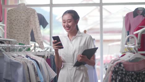 asian female owner of clothing store using smartphone while holding a tablet and walking around to check stock on rails